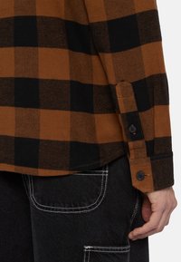 Brun og sort ternet flannel skjorte med knapærmer og afrundet kant; sat sammen med mørke cowboybukser med hvid syning.