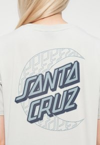 Hele hall hõbedane T-särk, millel on suur "SANTA CRUZ" graafika tumesinisel taustal, ümbritsetud ringikujulise, tsüklilise mustriga. Pehme kangast tekstuur.