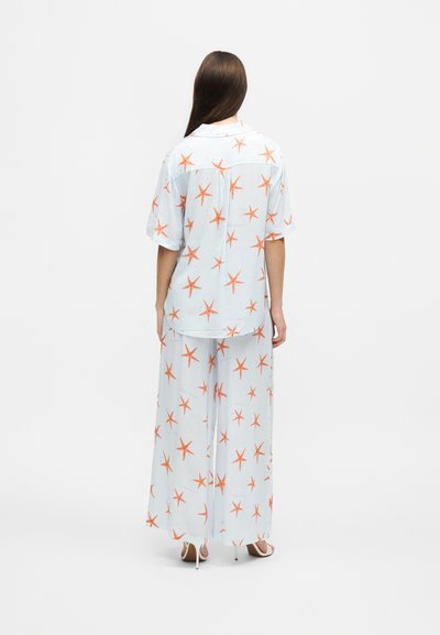 Femme debout de dos, portant un pantalon bleu clair à jambes larges et une chemise à manches courtes avec un motif d’étoiles de mer orange et des talons blancs.