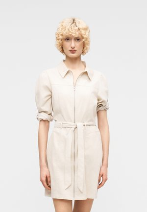 Moteris su trumpais garbanotais blondiniais plaukais, vilkinti smėlio spalvos su diržu suknele su susuktais rankovėliais ir priekyje esančiu užtrauktuku, stovinti priešais paprastą foną.