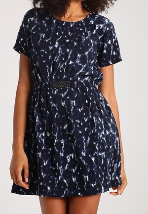 Femme portant une robe bleu marine à manches courtes et longueur genou, ornée de motifs abstraits blancs, ceinturée à la taille d'une ceinture noire tressée.