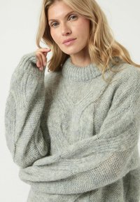 Jasnoszary sweter dziergany z wysokim, prążkowanym kołnierzem oraz luźnymi, oversize'owymi rękawami z wzorem w warkocz. Miękka i teksturowana tkanina.