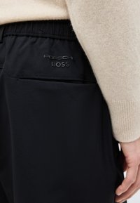 Κοντινό πλάνο στην τσέπη μαύρου παντελονιού με ελαστική μέση που δείχνει το λογότυπο "Porsche BOSS", δίπλα σε μπέζ πουλόβερ μακρυμάνικο και ένα χέρι.