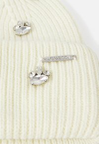 Berretto in maglia crema con costine verticali, caratterizzato da tre strass decorativi a forma di zampe e un'etichetta in argento con il logo "GUESS".