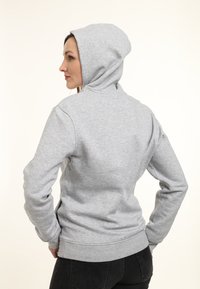 Felpa grigia con cappuccio, maniche lunghe e orlo a costine. Tessuto di cotone morbido con una texture liscia. Vista posteriore, il modello ha le mani sui fianchi.