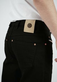 Svarta denimjeans med en slät textur, kopparnitar och en beige läderlapp med präglad logotyp på bakdelen av midjan.