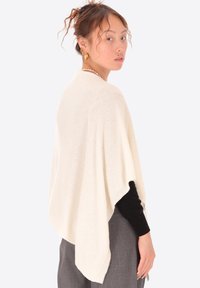 Poncho lavorato a maglia avorio con un design morbido e drappeggiato. Texture morbida e maniche larghe, indossato sopra un top nero e pantaloni grigi. Linee semplici e pulite.