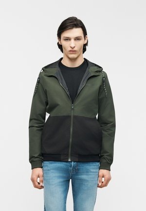 Mladý muž oblečený v zelenej a čiernej bunde na zips s kapucňou, pod ktorou má čierne tričko a modré džínsy, stojaci na jednoduchom pozadí.