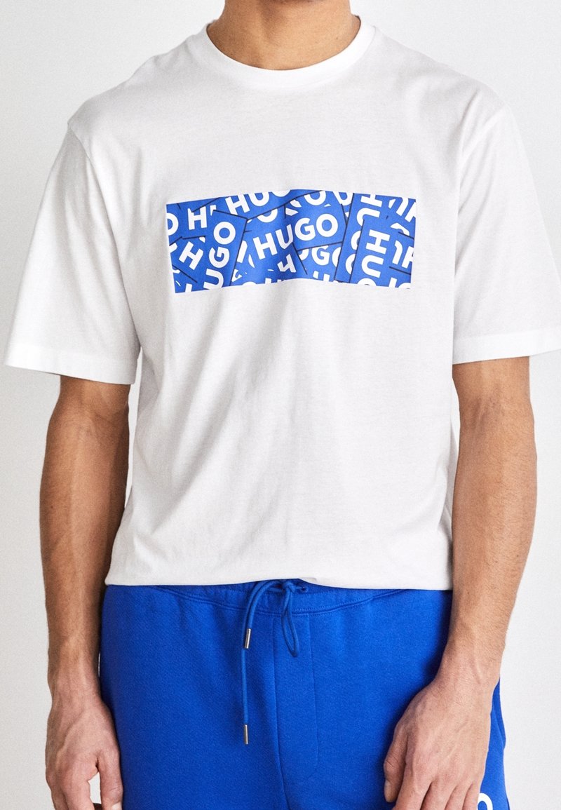 T-shirt en coton blanc avec un graphique rectangulaire bleu proéminent affichant le texte "HUGO" répété en blanc. Manches courtes et col rond.