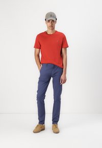 Tricou roșu cu mânecă scurtă și logo, pantaloni albaștri și adidași bej deschis. O șapcă gri completează ținuta. Design simplu și casual.