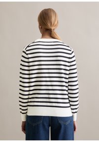 Personne aux cheveux blonds coiffés en queue de cheval basse, portant un pull à rayures horizontales blanches et noires à manches longues et un jean bleu, vue de dos.