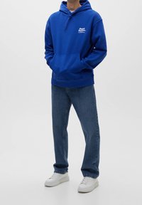 Blå huvtröja med känguruficka och snörning, med vit textlogotyp. Kombinerad med blå jeans och vita sneakers.