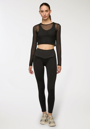 Top sportivo nero corto con maniche trasparenti, abbinato a leggings a vita alta. Tessuto liscio con design aderente e senza fantasie.