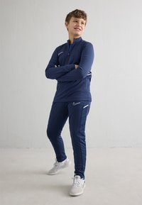 Marineblaues, langärmeliges Sportoberteil mit Reißverschluss und weißen Akzenten, kombiniert mit passenden marineblauen Hosen. Getragen mit grauen und weißen Turnschuhen. Glatte Textur.