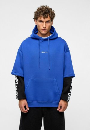 Blauwe oversized hoodie van zacht materiaal, met een kangoeroezak en witte tekst "IMPACT". Gelaagd met een zwart T-shirt met lange mouwen eronder.