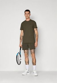 T-shirt de manga curta verde oliva e calções combinando, feitos de um material confortável, com tatuagens visíveis e uma raquete de ténis na mão.