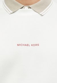 Λευκό πόλο μπλουζάκι με μπεζ γιακά που έχει λευκές ρίγες. Εμφανίζει το "MICHAEL KORS" με ροζ γραμματοσειρά στο μπροστινό μέρος. Λείο υφή.