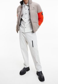 Bezešvá puffer bunda v béžové a oranžové barvě s vysokým límcem, se zipem vpředu a detaily loga. V kombinaci s bílými kalhotami a černými platformovými botami.