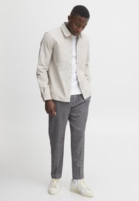 Lys beige skjorte med knappestolpe og to brystlommer, båret over en hvit t-skjorte, kombinert med grå teksturerte bukser og hvite sneakers.
