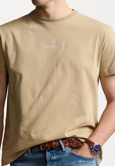 Tricou bej cu mânecă scurtă și guler cu nervuri, având un logo brodat alb "Polo Ralph Lauren" pe piept. Purtați cu blugi albaștri.