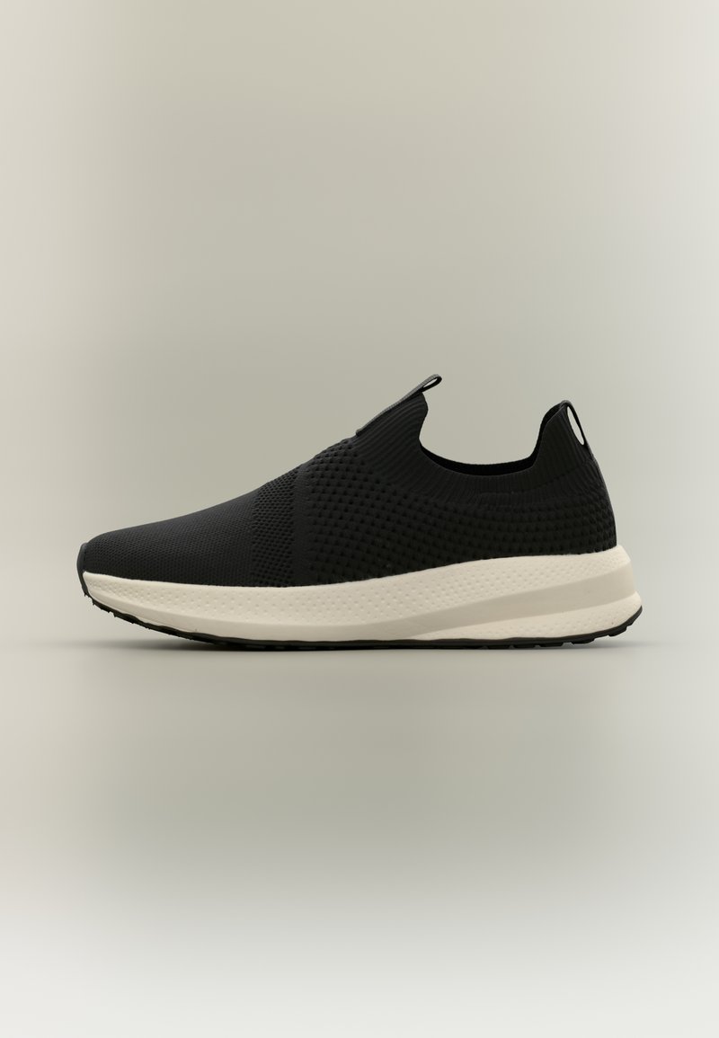 Μαύρο πλεκτό αθλητικό παπούτσι slip-on με υφασμάτινο επάνω μέρος με υφή και χοντρή λευκή μαλακή σόλα σε ουδέτερο φόντο.