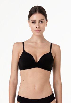 Donna con capelli scuri raccolti che indossa un reggiseno nero e mutandine nere abbinate su uno sfondo bianco uniforme.