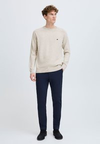 Beige strikkegenser med rund hals, kombinert med marineblå slim-fit bukser. Svart ankelstøvler og en liten logodetalj på genseren.