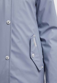 Veste imperméable gris clair avec boutons-pression, dotée d'une poche diagonale zippée et d'une texture lisse. Le tissu est légèrement réfléchissant.