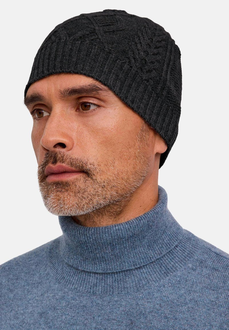 Hombre con barba recortada que lleva un gorro de punto gris oscuro y un suéter de cuello alto azul, mirando ligeramente hacia un lado.