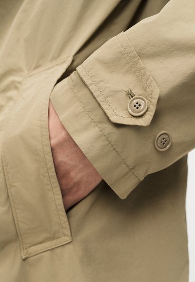 Χέρι που εισέρχεται στην τσέπη μπεζ παλτού, μανίκι παλτού με δύο μπεζ κουμπιά ορατά, κοντινή λεπτομέρεια υφάσματος και ραφής.