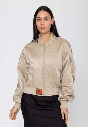 Donna con una giacca bomber beige con tasche con zip e una gonna nera, in piedi davanti a uno sfondo chiaro e uniforme.