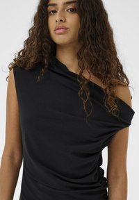 Ung kvinde med krøllet mørkt hår iført en one-shoulder sort top foran en ensfarvet lys baggrund.