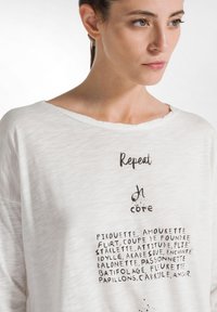 Weißes, oversized Hemd aus leichtem, strukturiertem Stoff mit schwarzem Drucktext in verschiedenen Schriftarten und -größen. Kurzer, weiter Ausschnitt.