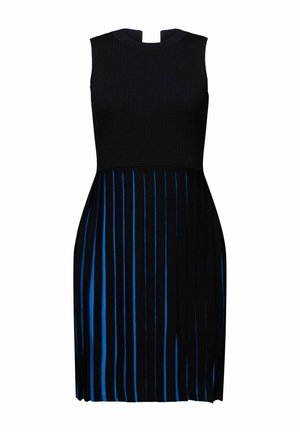 Femme portant une jupe rayée noire et bleue, un haut noir, des chaussettes bleu marine, des chaussures noires, avec une écharpe jaune et tenant une veste bleu marine sur un bras.