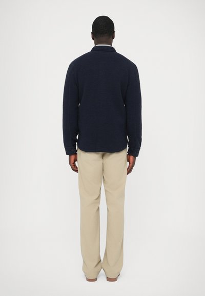 Temno modra flis pulover z teksturirano površino, stoječim ovratnikom in dolgimi rokavi, kombiniran z bež, ohlapnimi hlačami. Zadnji pogled.