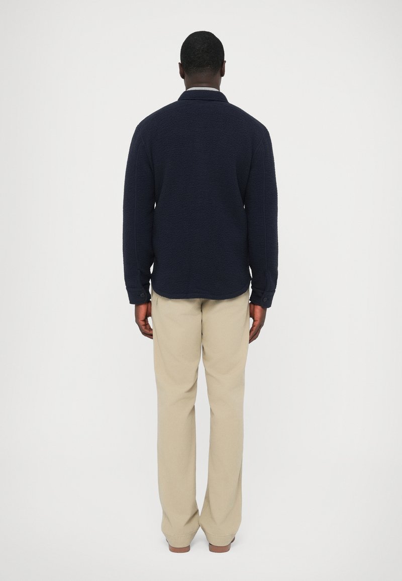 Navy fleece pullover with a textured surface, a stand-up collar, and long sleeves paired with beige, loose-fitting trousers. Back view.