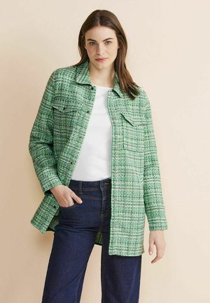 Chaqueta de tweed verde con cierre de botón, dos bolsillos en el pecho y un corte relajado, combinada con una camiseta blanca y jeans oscuros.