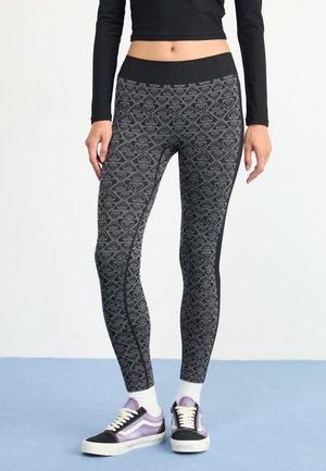 Leggings con diseño en negro y gris, que cuentan con una cinturilla ancha, un diseño geométrico y un corte ajustado, combinadas con zapatillas negras y moradas.