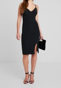 Eng anliegendes schwarzes Kleid mit tiefem V-Ausschnitt, dünnen Trägern und einem Schlitz bis zum Oberschenkel. Kombiniert mit schwarzen Riemchenabsätzen und einer Clutch in der Hand.