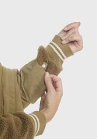 Braune Jacke mit strukturiertem, sherpa-gefüttertem Bund; elastischer Strickbund mit weißen Streifen. Eine Hand stellt den Druckknopfverschluss des Bundes ein.