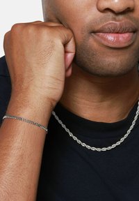 Collana in argento a catena attorcigliata e bracciale sottile indossati su un polso, su una camicia nera. Texture liscia, finitura riflettente, design minimalista.