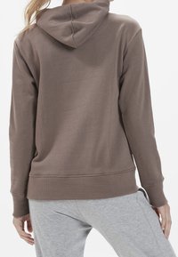 Felpa marrone in cotone con vestibilità comoda, tasca a marsupio e polsini a coste. Sotto, sono indossati pantaloni della tuta grigi che mostrano una texture morbida.