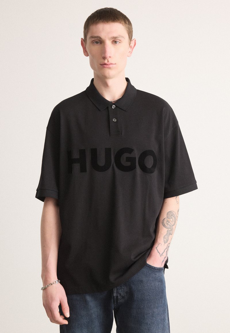 Juodas polo marškinėliai su trimis mygtukais ant apykaklės, centre iškilus didelis "HUGO" logotipas. Trumpomis rankovėmis, laisva forma.