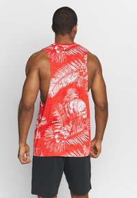 Top senza maniche rosso con un motivo di foglie di palma bianche, realizzato in tessuto leggero. Il retro presenta un design con taglio aperto. Indossati pantaloni corti neri.