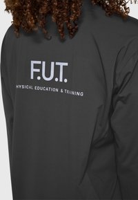 Czarna sportowa kurtka z nadrukiem "F.U.T." oraz "PHYSICAL EDUCATION & TRAINING" w białym kolorze na plecach. Gładka tekstura, dopasowany fason.