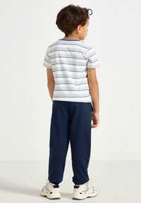 Valkoisessa, sinisessä ja oranssissa raidallisessa puuvillaisessa T-paidassa; tummansininen collegehousut; kumi-pohjaiset tennarit; lapsi kuvattuna takaa.