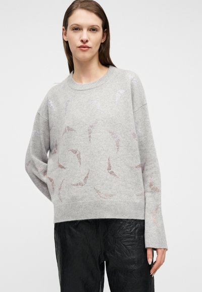 Γυναίκα που φοράει ένα ανοιχτό γκρι πουλόβερ με διακριτικά μεταλλικά σχέδια πουλιών και υφασμένα μαύρα παντελόνια, στέκεται μπροστά σε απλό ανοιχτό φόντο.