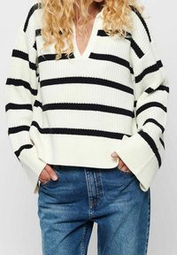 Gestreifter Oversized-Pullover in Schwarz und Weiß mit V-Ausschnitt, gerippter Textur und weiten Ärmeln, kombiniert mit blauen Jeans.