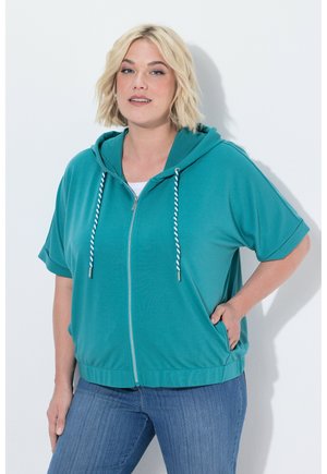 Lyshåret kvinde iført teal lynlåshoodie med hvide snore og blå jeans, stående med den ene hånd i lommen mod hvid baggrund.