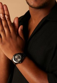 Man in zwart shirt met handen tegen elkaar gedrukt, draagt een zwart horloge met zilveren en rode details aan de linkerpols.